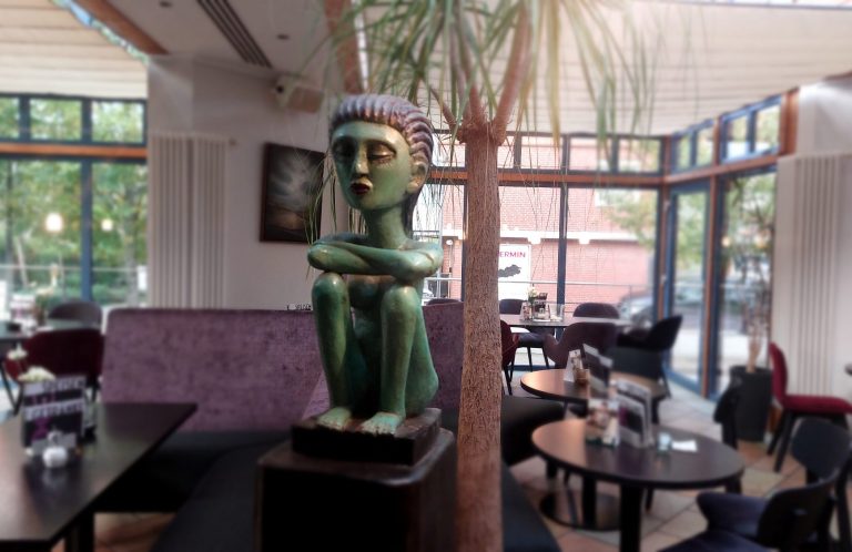 Grüne Statue einer sitzenden Figur in einem Café mit Palmen und modernen Möbeln.
